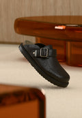 Schwarzer Slip-On-Clog aus Leder mit Schnalle und durchlochtestem Muster, auf hellem Teppich neben einem durchscheinenden braunen Tisch platziert.