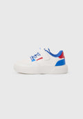 Weißer Sneaker mit blauen und roten Akzenten, blauen Schnürsenkeln und einem Klettverschluss, von der Seite auf einfarbigem Hintergrund gezeigt.