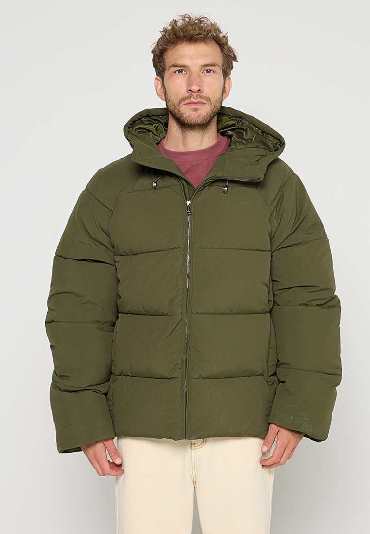 Mann in olivgrüner Daunenjacke mit Kapuze, maroonem Shirt und hellen Hosen, stehend vor einem schlichten Hintergrund.