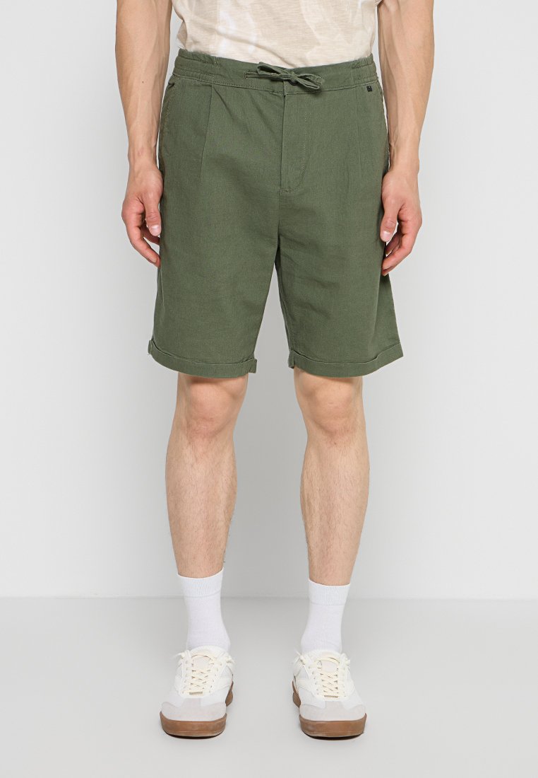 Mann trägt olivgrüne knielange Shorts mit umgeschlagenem Saum, weiße Socken und weiße Sneakers, steht vor einfarbigem Hintergrund.