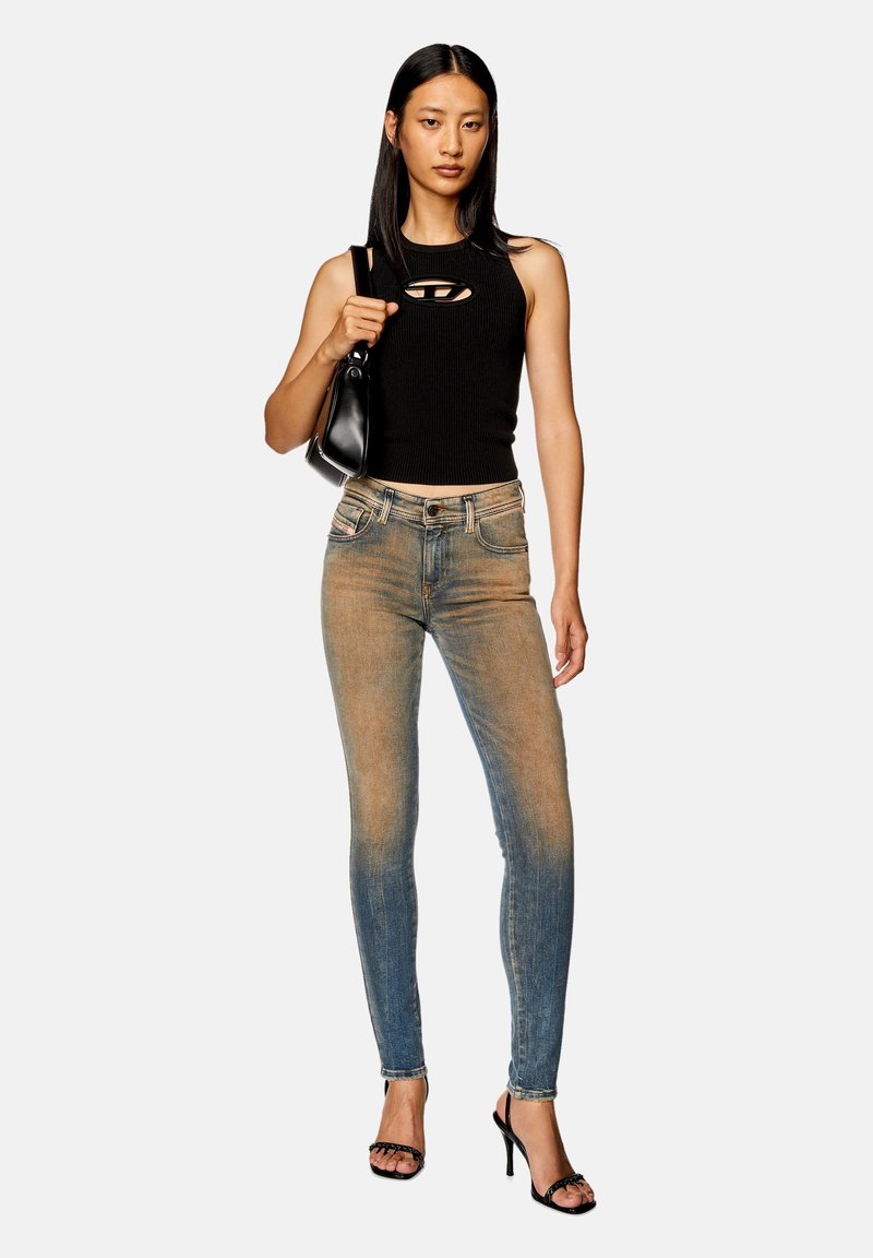 Schwarzes, geripptes, ärmelloses Oberteil mit Ausschnitt, kombiniert mit gefädelten blauen Skinny Jeans und schwarzen, geschnürten High Heels. Model hält eine schwarze Tasche.