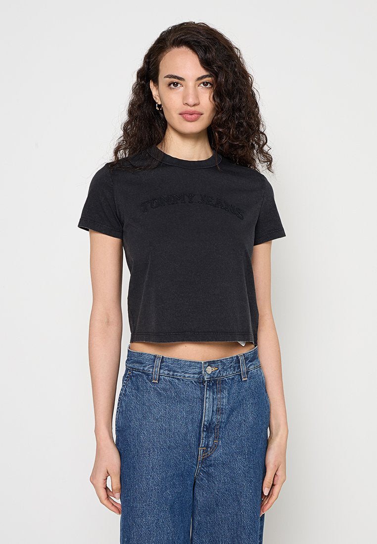 Frau mit lockigem dunklem Haar trägt ein schwarzes, kurzes Tommy Jeans T-Shirt und hoch taillierte blaue Jeans und steht vor einem schlichten hellen Hintergrund.