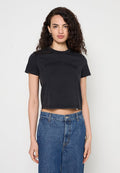 Frau mit lockigem dunklem Haar trägt ein schwarzes, kurzes Tommy Jeans T-Shirt und hoch taillierte blaue Jeans und steht vor einem schlichten hellen Hintergrund.