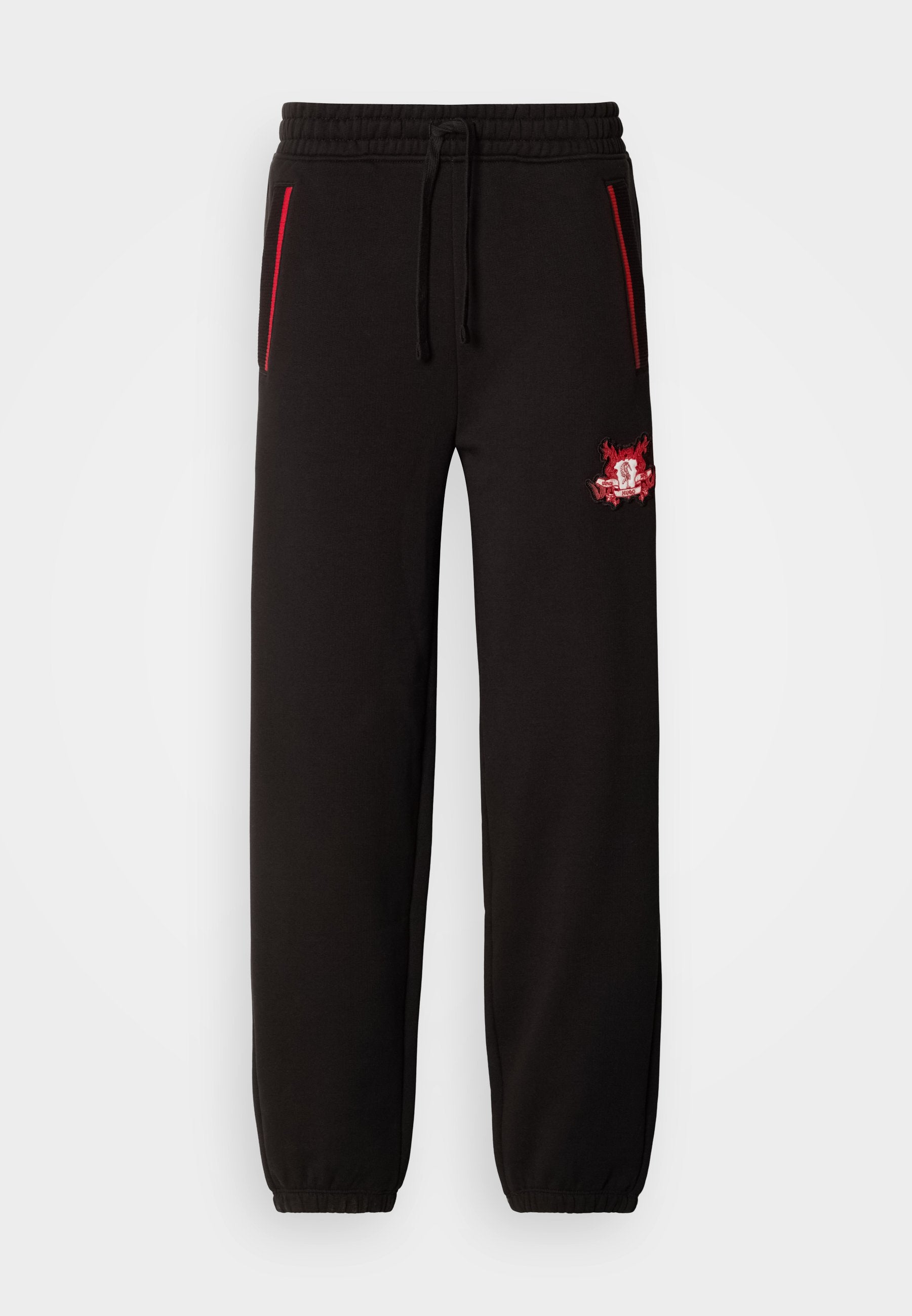 Schwarze Jogginghose mit elastischem Bund, roten Akzentreißverschlüssen und einem auffälligen gestickten Logo auf dem linken Oberschenkel. Glatte Textur, schmaler Schnitt.