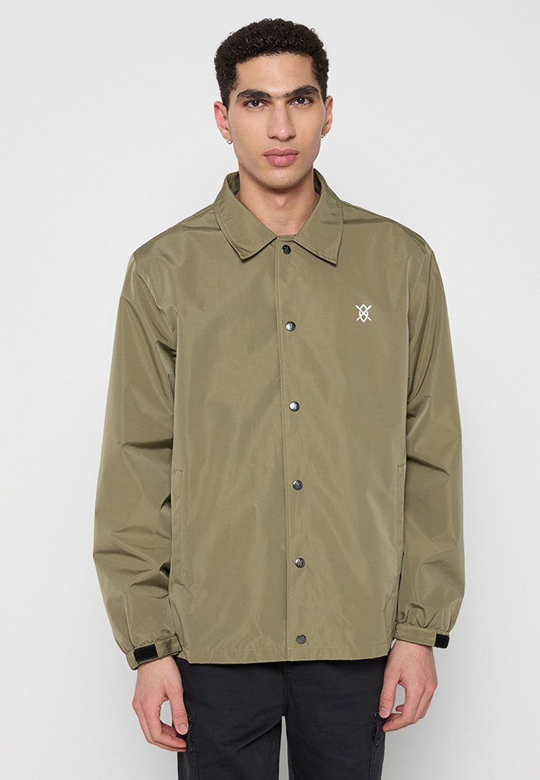Junger Mann in olivgrüner, knöpfbarer Jacke mit kleinem weißen Logo auf der Brust, steht vor einem schlichten hellen Hintergrund.