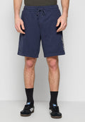 Mann in dunkelblauen Joggingshorts mit Kordelzug, schwarzen Socken und schwarzen Adidas-Sneakern mit weißen Streifen, der gegen eine schlichte helle Wand steht.