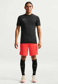 Mann steht und trägt ein schwarzes Nike-Sportshirt, rote Shorts, schwarze Socken und weiße Stollenschuhe vor einem schlichten weißen Hintergrund.