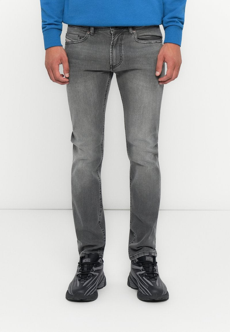 Mann in verblichenen grauen Slim-Fit-Jeans, schwarzen und grauen Diesel-Sneakern und einem blauen Langarmoberteil vor einem schlichten weißen Hintergrund.