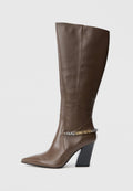 Braune Lederstiefel bis zum Knie mit spitzer Zehe, dicker schwarzer Absatz und metallischer Kettenakzent um den Knöchel. Glatte Textur, minimales Design.