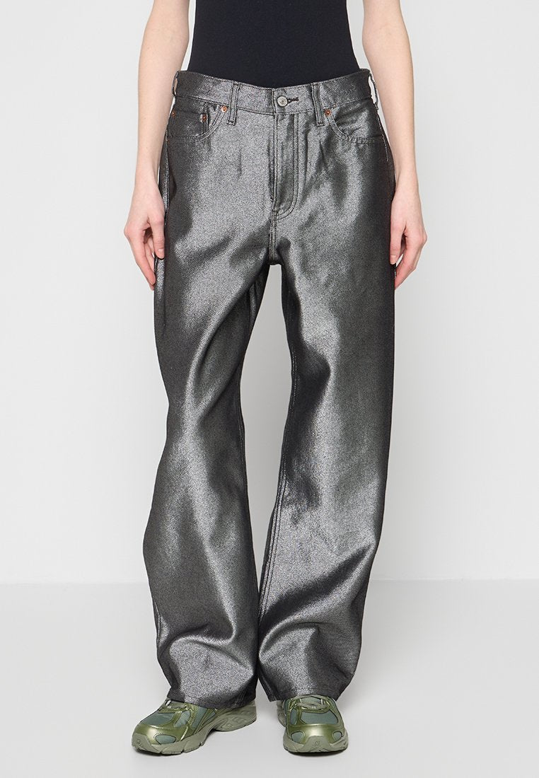 Person trägt metallisch silberne weite Hose mit Fronttaschen und grüne Sportschuhe und steht vor einem einfarbigen Hintergrund.