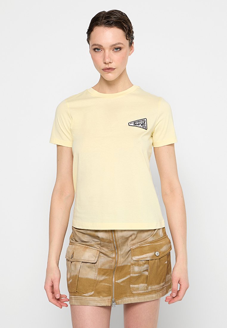 Junge Frau in einem blassgelben kurzärmeligen Shirt mit kleinem Frontlogo und einem beigen Batik-Minirock mit Reißverschluss und Taschen.