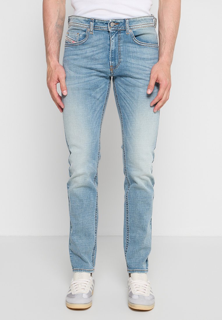 Mann trägt hellblaue Slim-Fit-Jeans und weiße Sneakers und steht vor einem einfarbigen Hintergrund mit entspannt an den Seiten hängenden Händen.