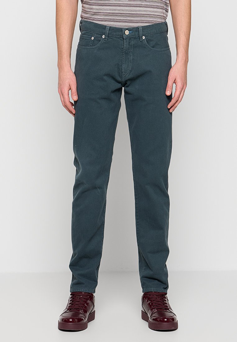 Eng anliegende grüne Herrenhose mit glatter Textur, Fünf-Taschen-Design, Metallknopfverschluss und kombiniert mit burgunderfarbenen Sneakern.