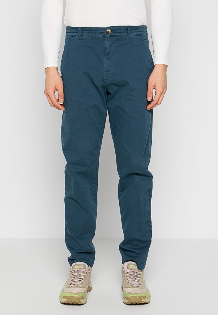 Herren Chinos aus blauer Baumwolle mit geradem Bein, Knopfverschluss, Vordertaschen und glatter Textur, kombiniert mit beigen Sneakers.