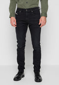 Mann in schwarzen Slim-Fit-Jeans, schwarzen Lederschuhen und einem olivgrünen, knopfgebundenen Hemd, der vor einem schlichten Hintergrund steht.