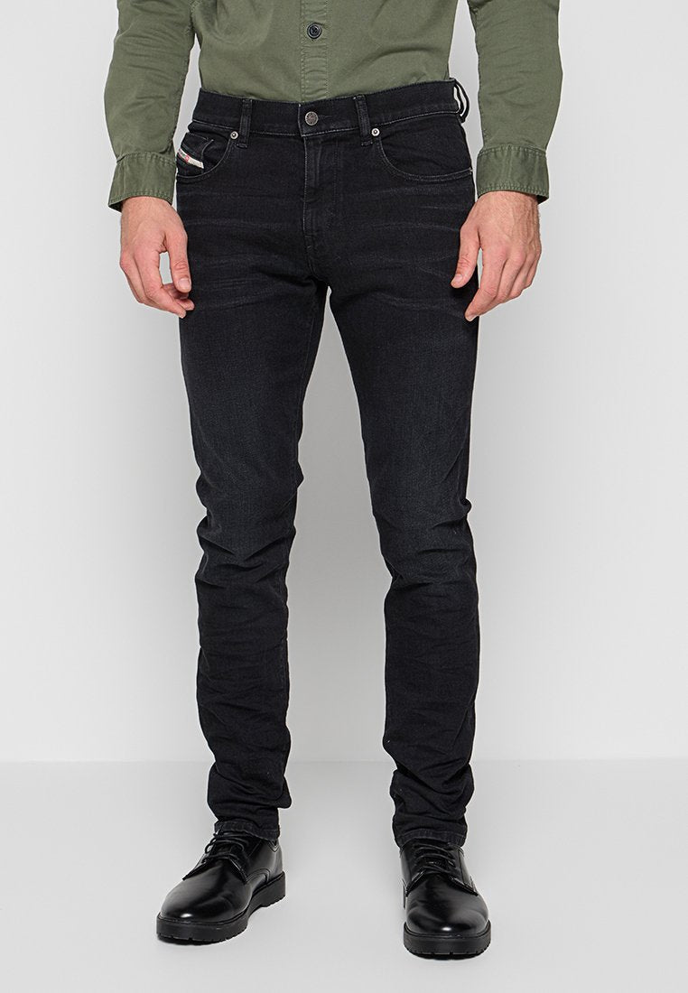 Mann in schwarzen Slim-Fit-Jeans, schwarzen Lederschuhen und einem olivgrünen, knopfgebundenen Hemd, der vor einem schlichten Hintergrund steht.