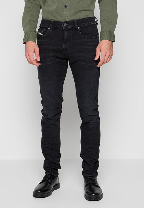 Mann in schwarzen Slim-Fit-Jeans, schwarzen Lederschuhen und einem olivgrünen, knopfgebundenen Hemd, der vor einem schlichten Hintergrund steht.