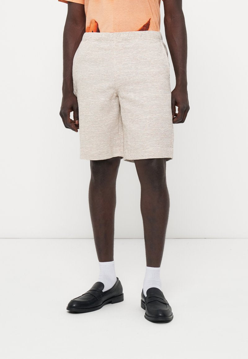 Mann trägt beige gestrickte Shorts, schwarze Loafers, weiße Socken und ein oranges T-Shirt mit einem teilweisen Karottengrafik.