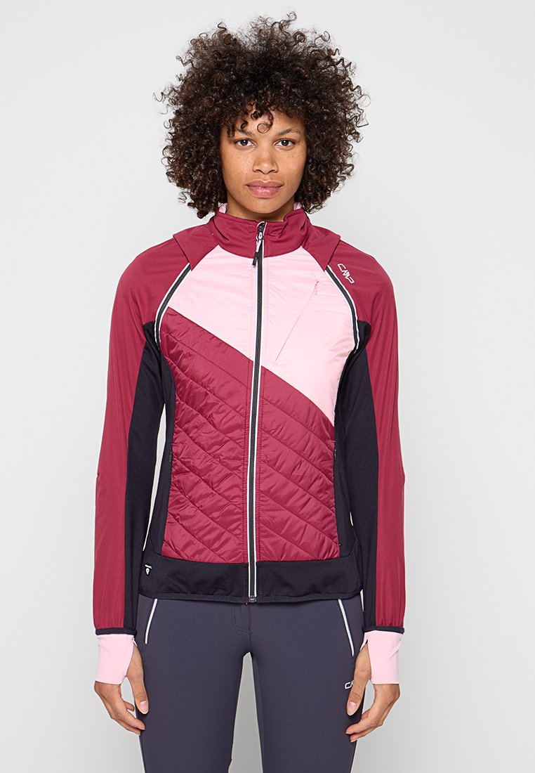 Frau mit lockigem Haar, die eine sportliche Jacke mit Reißverschluss mit bordeauxroten, pinken und schwarzen Einsätzen trägt, vor einem schlichten hellen Hintergrund stehend.