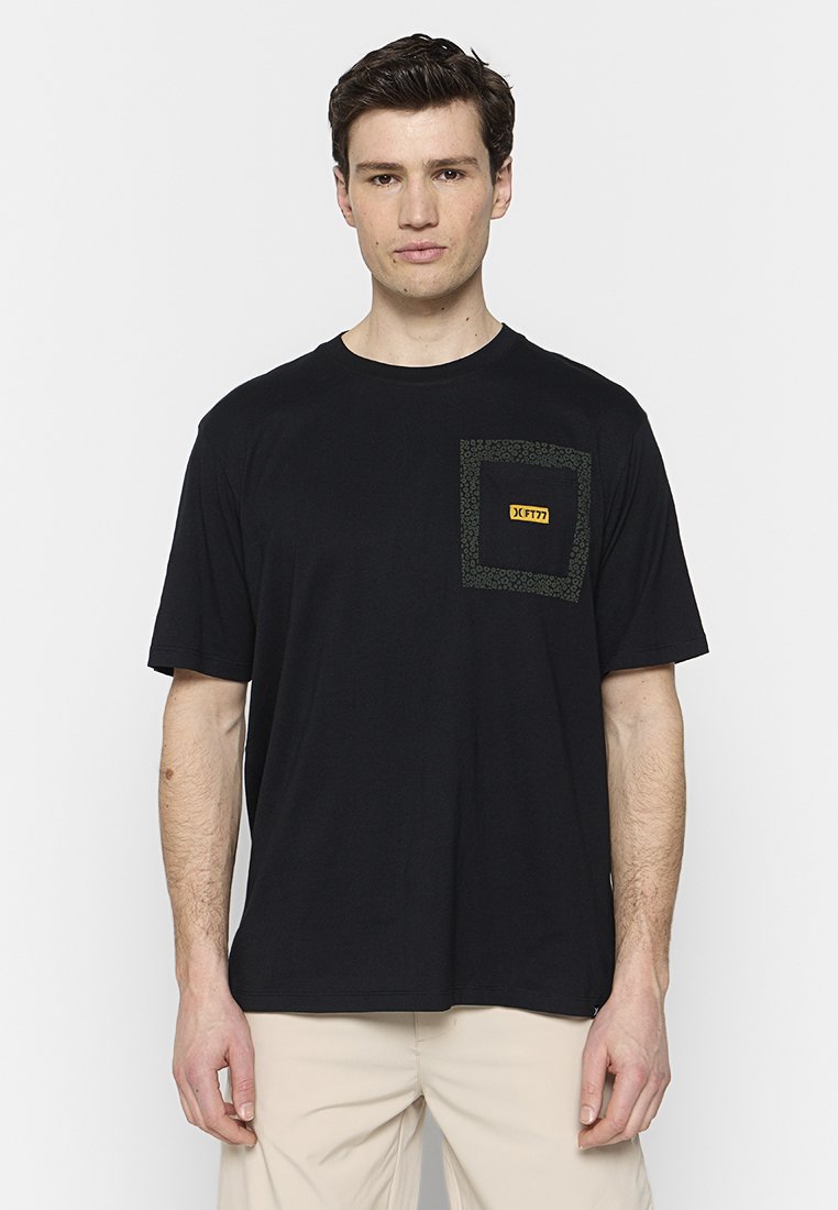 Schwarzes T-Shirt mit kurzen Ärmeln aus Baumwolle, ausgestattet mit einer grünen gemusterten Tasche und einem gelben Logosticker auf der Brust. Reguläre Passform.