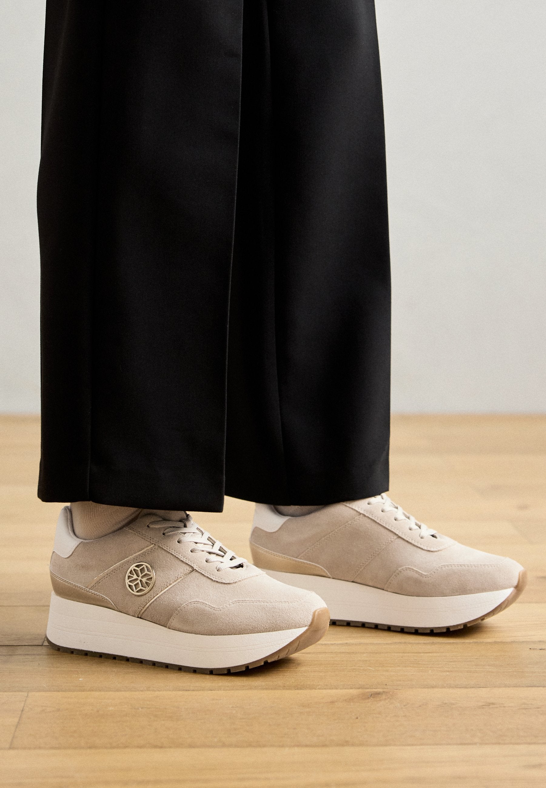 Beige Wildlederschuhe mit einer weißen Plateausohle, ausgestattet mit einer Schnürdesign, geprägtem Logo und strukturierten Akzenten. Kombiniert mit schwarzen Hosen.