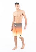 Junger Mann, der barfuß steht und orangefarbene Gradient-Bade shorts mit Kordelzug trägt, nach unten blickt und lächelt, vor einem weißen Hintergrund.
