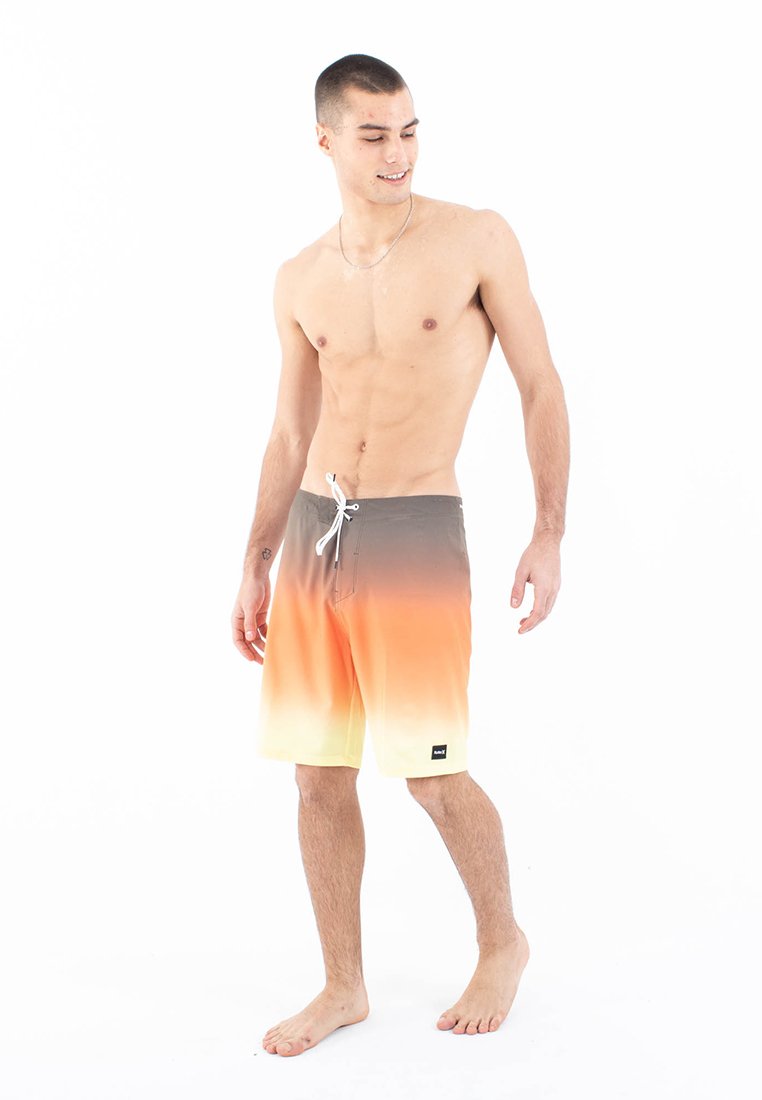Junger Mann, der barfuß steht und orangefarbene Gradient-Bade shorts mit Kordelzug trägt, nach unten blickt und lächelt, vor einem weißen Hintergrund.