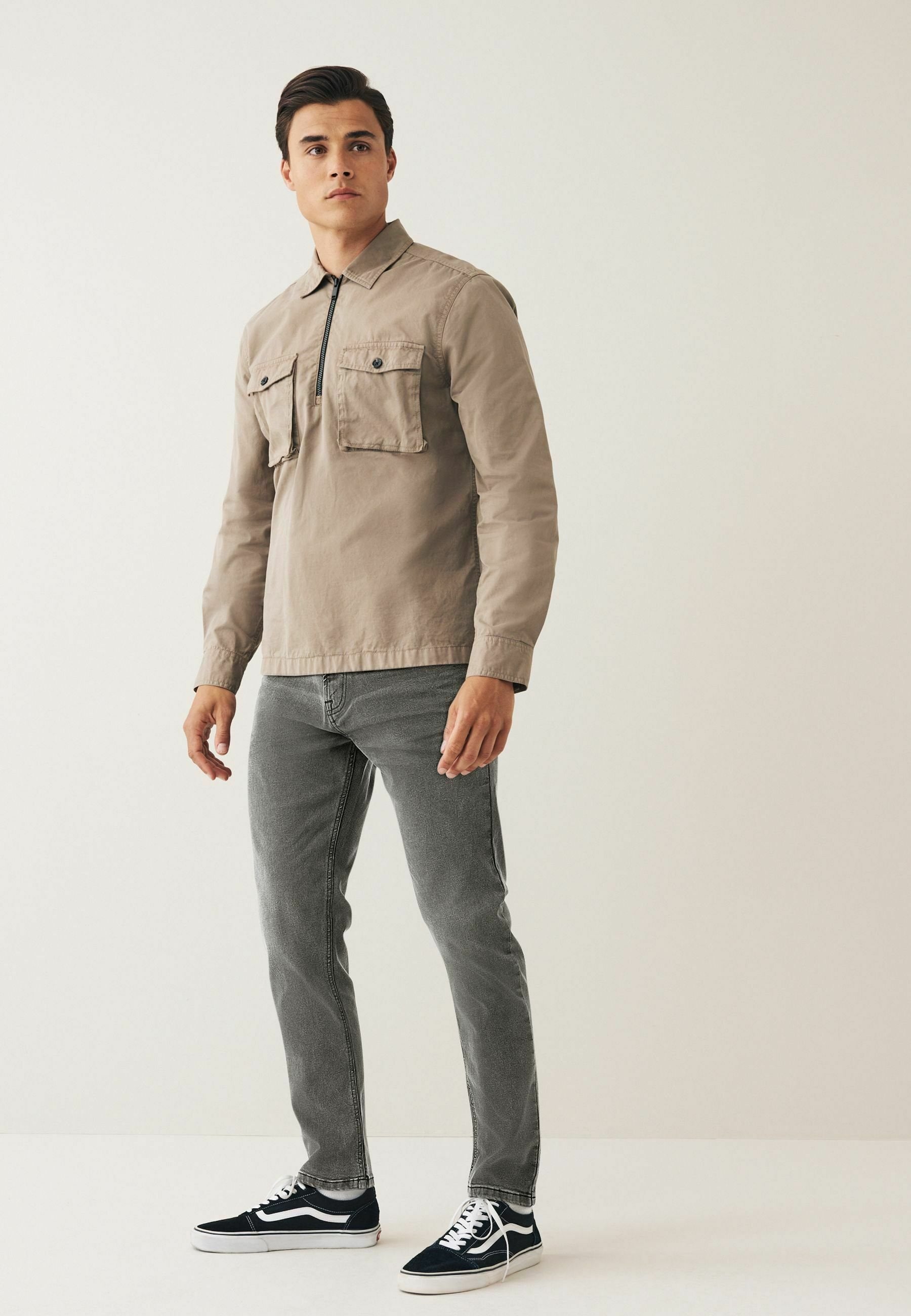 Hellbraunes Langarmhemd mit zwei Brusttaschen und einem Reißverschluss, kombiniert mit dunkelgrauen Jeans und schwarzen Sneakers, vor einem schlichten Hintergrund stehend.