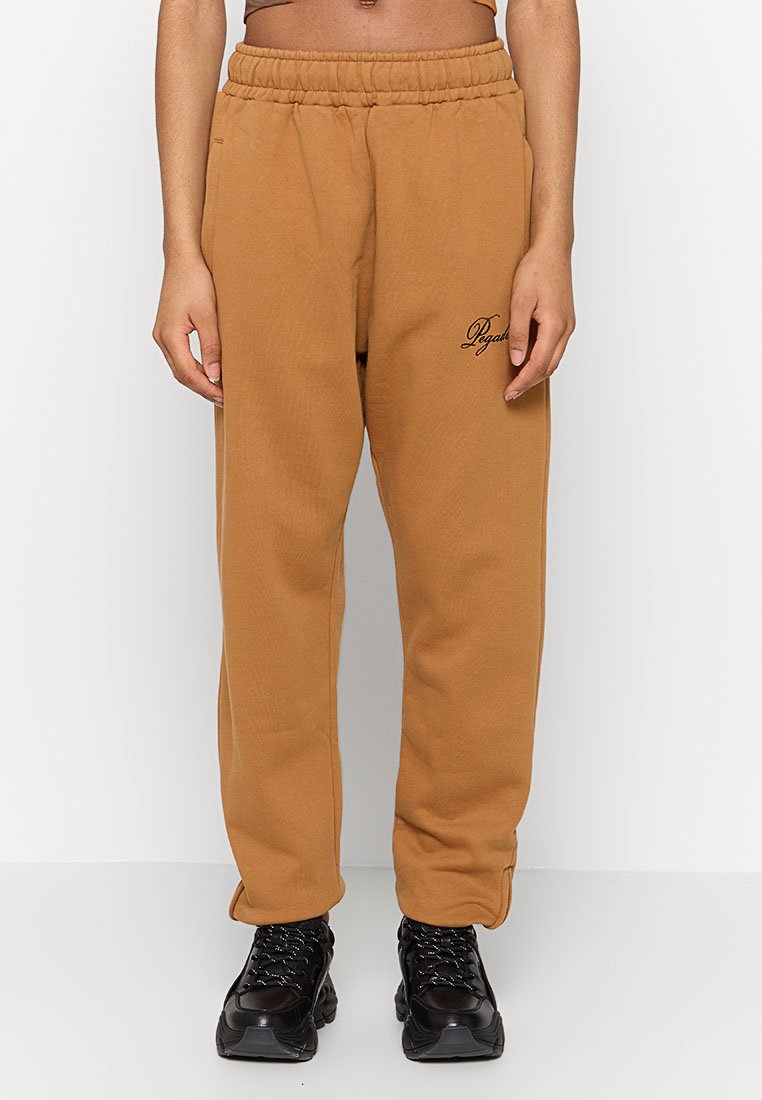 Braune Sweatpants mit elastischem Bund und seitlichen Taschen, kombiniert mit schwarzen klobigen Turnschuhen, getragen von einer Person, die vor einem weißen Hintergrund steht.