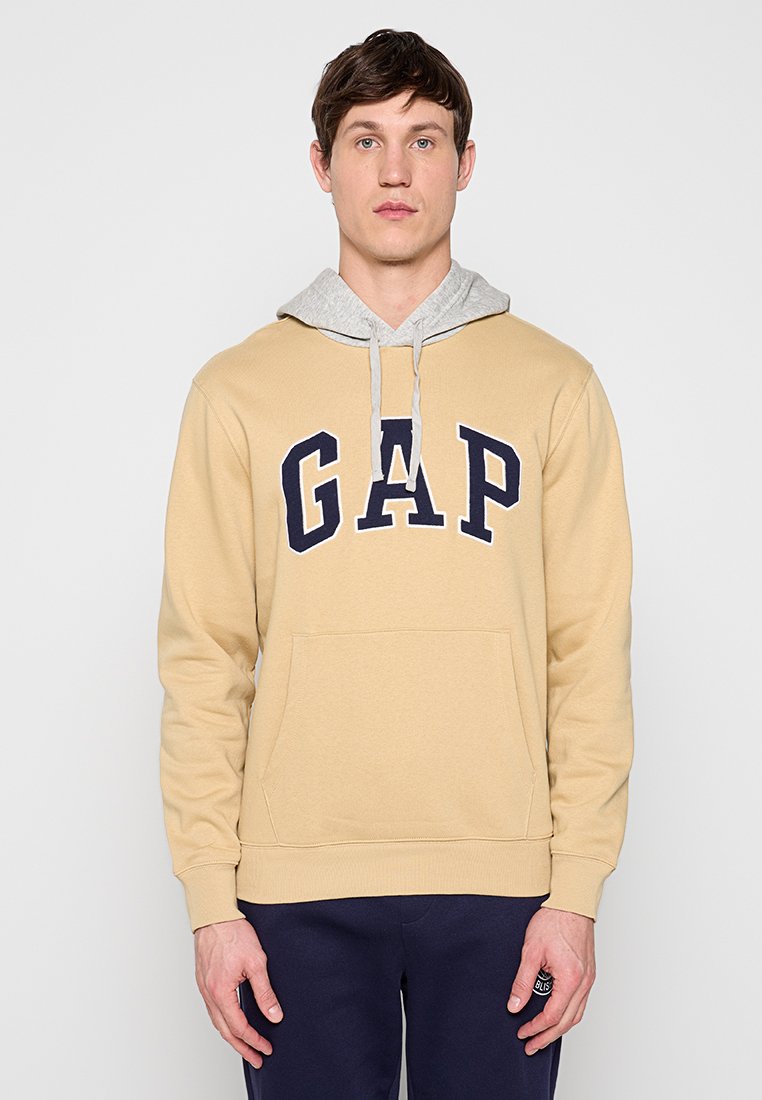 Mann trägt beige GAP-Hoodie mit grauer Kapuze und dunkelblauer Hose, steht frontal vor einem schlichten hellen Hintergrund.