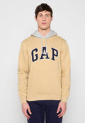 Mann trägt beige GAP-Hoodie mit grauer Kapuze und dunkelblauer Hose, steht frontal vor einem schlichten hellen Hintergrund.