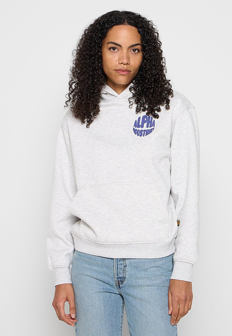 Frau mit lockigem Haar, die einen hellgrauen Alpha Industries Hoodie und hellblaue Jeans trägt, steht vor einem schlichten weißen Hintergrund.