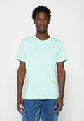 Mann mit kurzen lockigen Haaren, der ein hellmintgrünes T-Shirt und blaue Denim-Cargohosen trägt, steht vor einem schlichten weißen Hintergrund.
