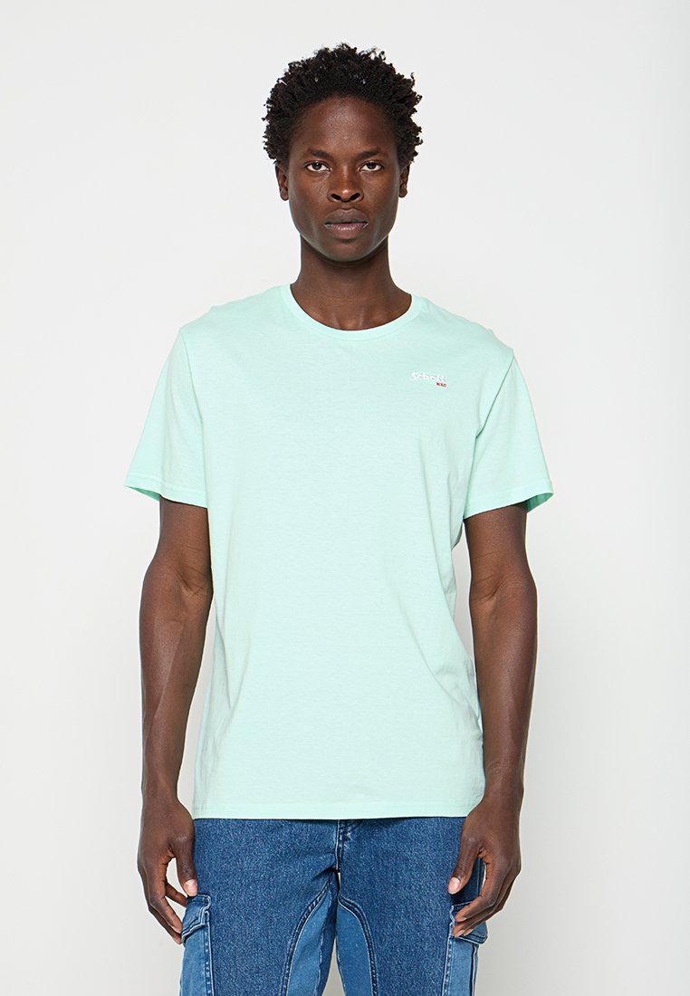 Mann mit kurzen lockigen Haaren, der ein hellmintgrünes T-Shirt und blaue Denim-Cargohosen trägt, steht vor einem schlichten weißen Hintergrund.