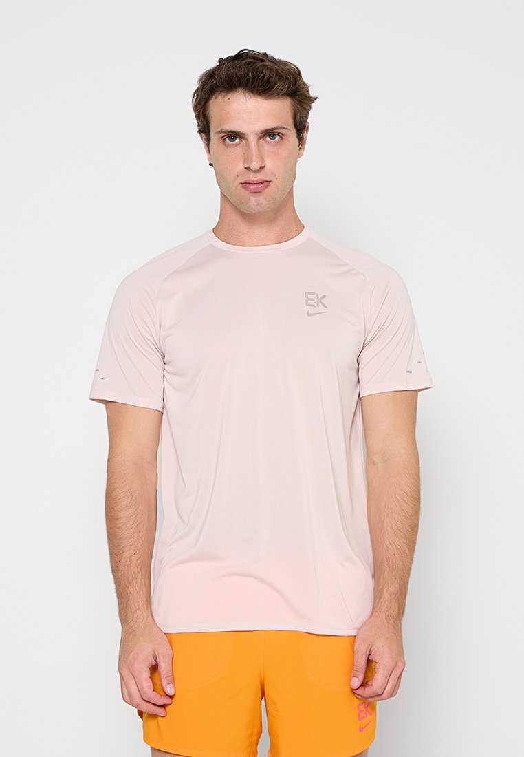 Mann trägt hellrosa kurzärmeliges Sportshirt und leuchtend orangefarbene Shorts und steht vor einem schlichten weißen Hintergrund.