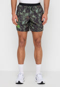 Mann trägt grüne und schwarze Shorts mit Blattmuster und schwarzem Bund, weiße Socken und weiße Laufschuhe, steht vor einfarbigem Hintergrund.