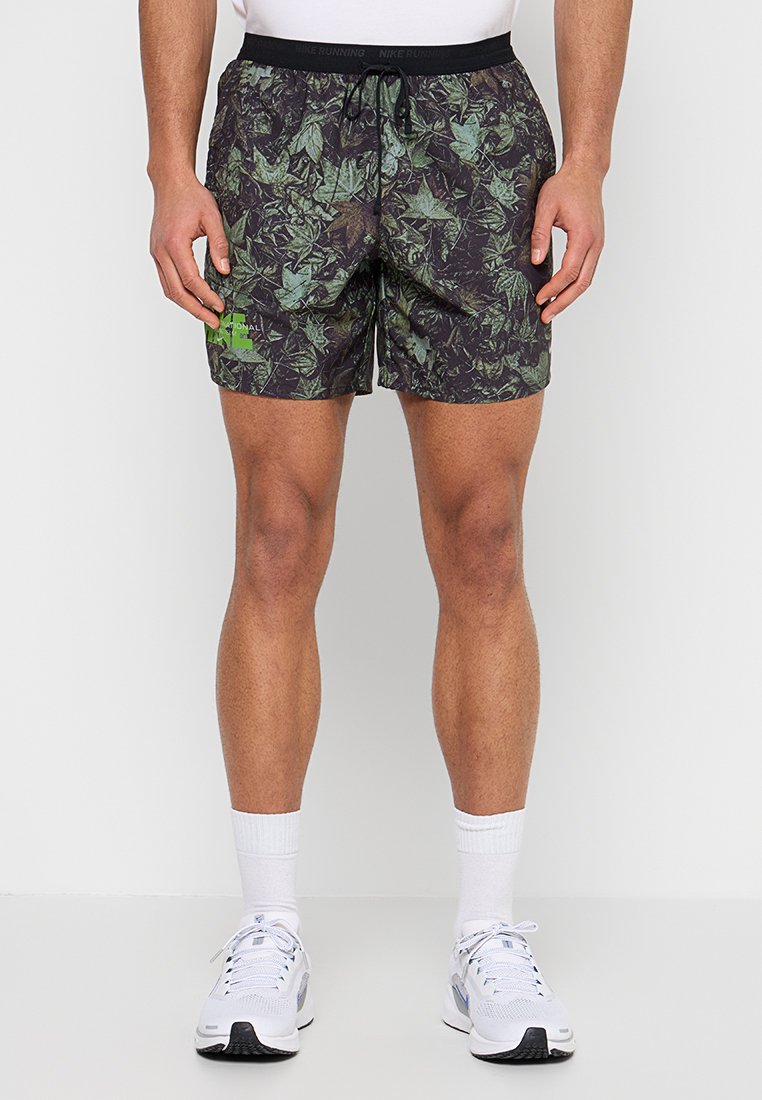 Mann trägt grüne und schwarze Shorts mit Blattmuster und schwarzem Bund, weiße Socken und weiße Laufschuhe, steht vor einfarbigem Hintergrund.