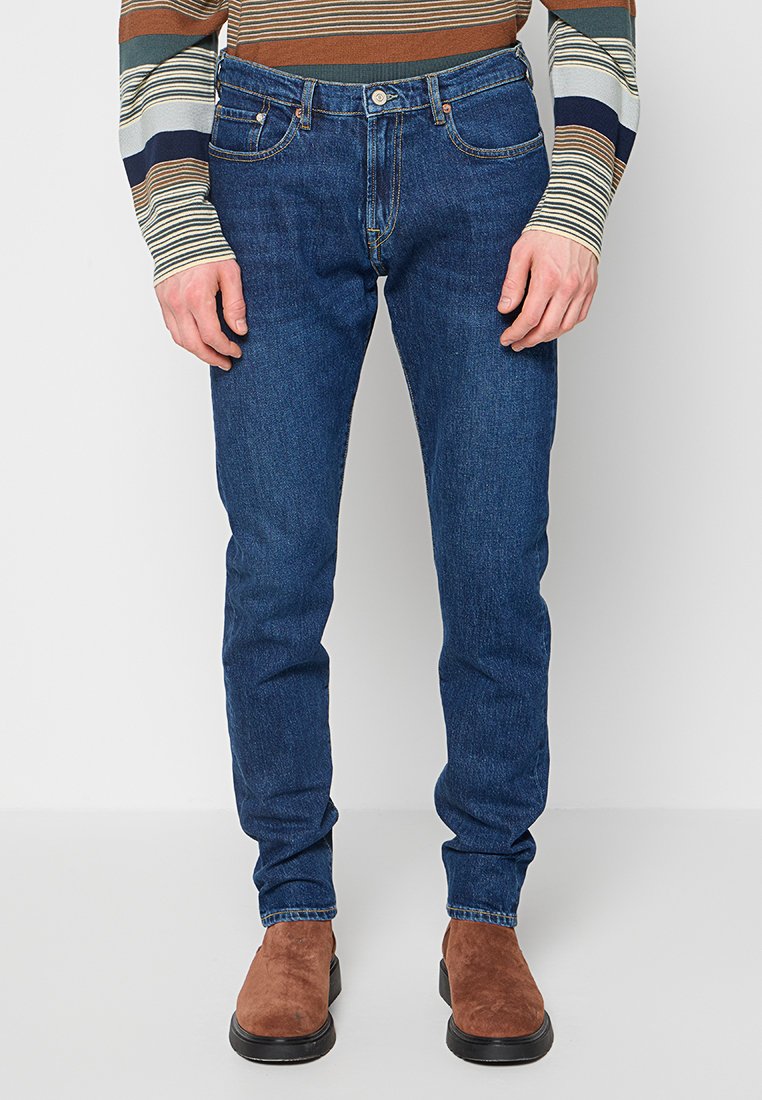 Dunkelblaue Denim-Jeans mit figurbetontem Schnitt, Fünf-Taschen-Stil und dezenten Kontrastnähten. Kombiniert mit braunen Wildleder-Stiefeletten.