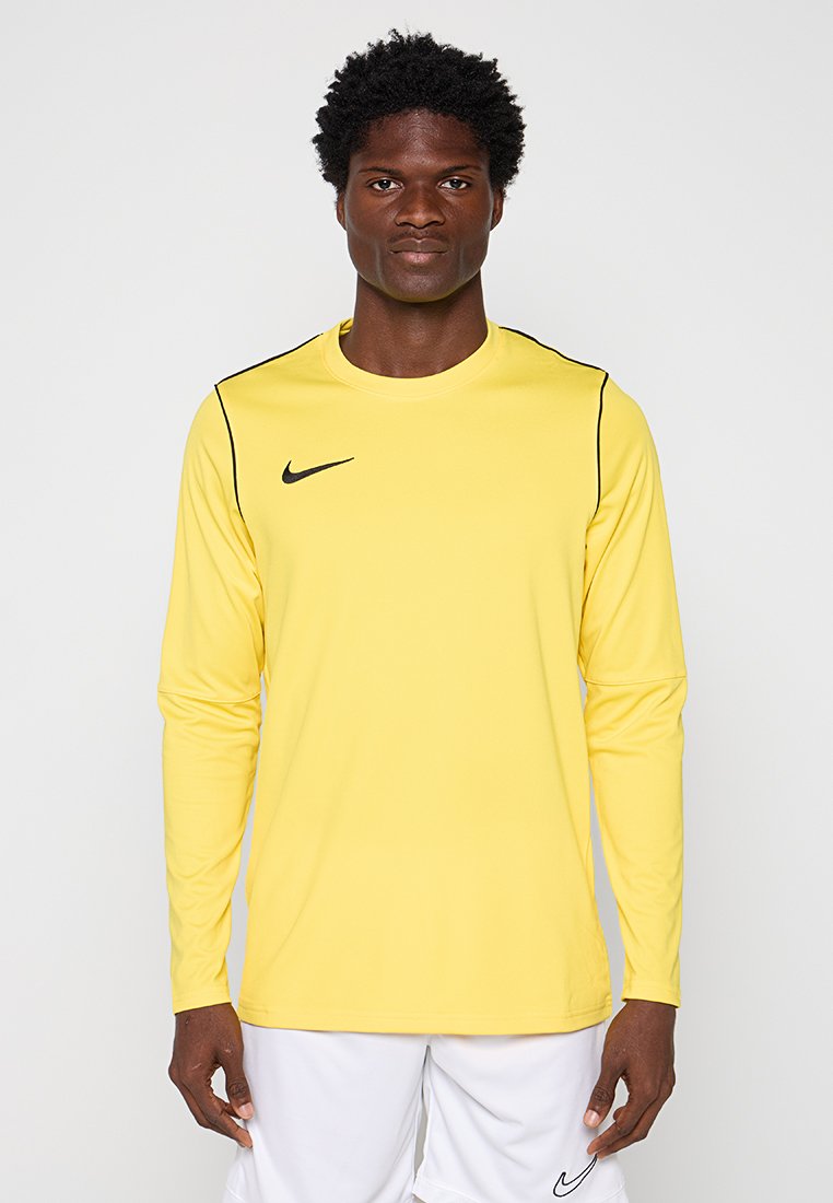 Junger Mann trägt ein gelbes langärmeliges Nike-Sportshirt und weiße Nike-Shorts, steht vor einem schlichten hellen Hintergrund.