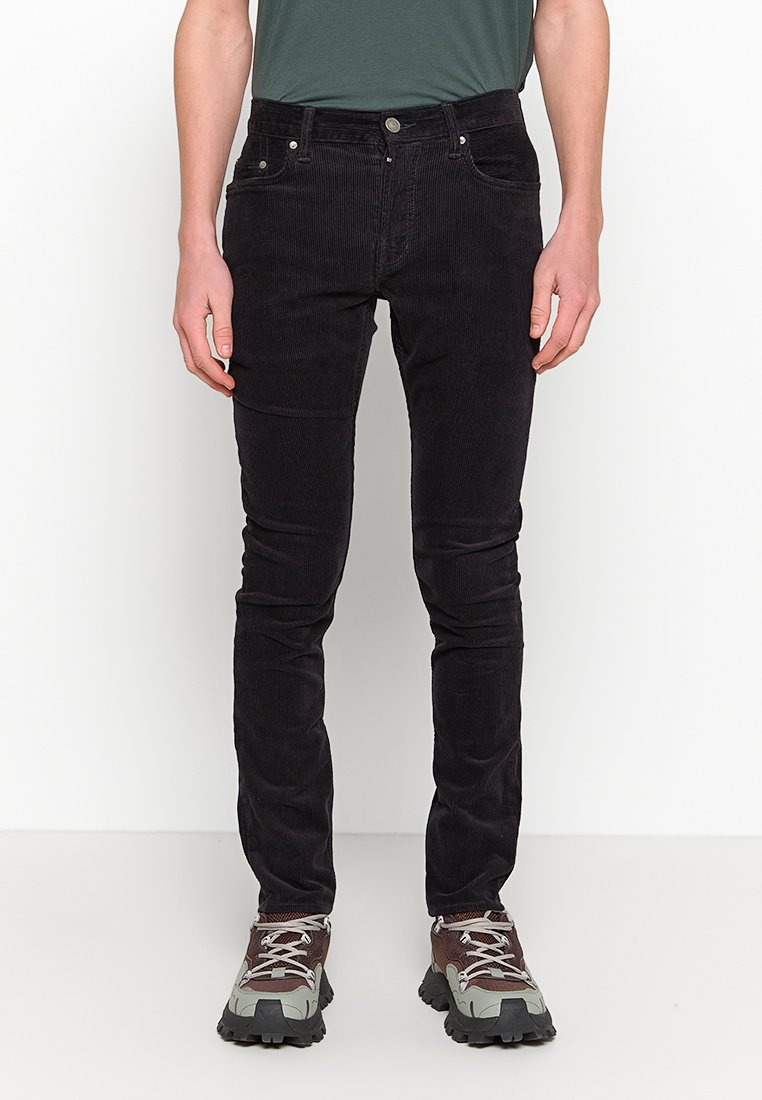 Schwarze Cordjeans im Slim-Fit, mit fünf Taschen, Nähtelementen und einem Reißverschluss. Kombiniert mit grauen und braunen Sneakers.