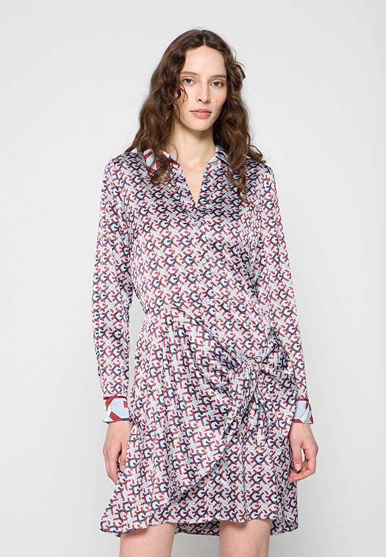 Frau mit langen, lockigen Haaren, die ein langärmliges, gemustertes Kleid mit roten, blauen und weißen geometrischen Formen und einer gerafften Taillendetail trägt.