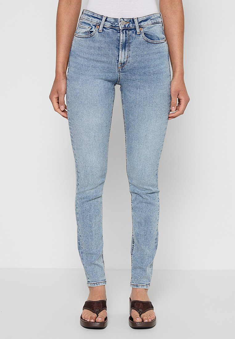 Frau trägt hellblaue Skinny-Jeans mit hoher Taille, kombiniert mit dunkelbraunen Plateausandalen, steht vor einem einfarbigen Hintergrund.