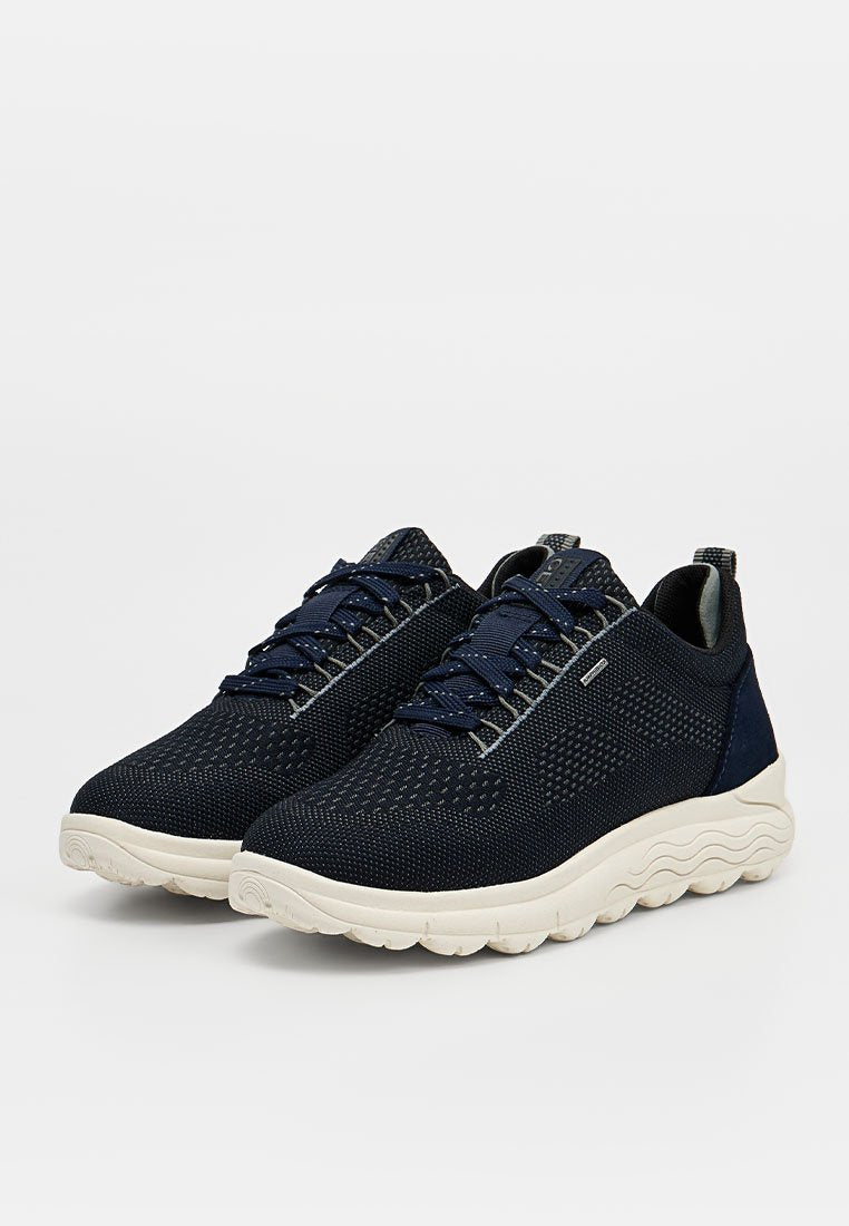 Paar marineblaue gestrickte Sneaker mit weißen wellenförmigen Sohlen, Schnürverschluss und Zuglaschen hinten vor einfarbigem Hintergrund.