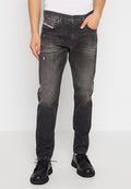 Mann in abgewetzten schwarzen Jeans mit Distressed-Effekten, schwarzen Schnürschuhen und einem weißen ärmellosen Oberteil, der mit den Händen in den Taschen steht.