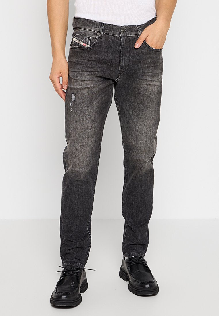 Mann in abgewetzten schwarzen Jeans mit Distressed-Effekten, schwarzen Schnürschuhen und einem weißen ärmellosen Oberteil, der mit den Händen in den Taschen steht.
