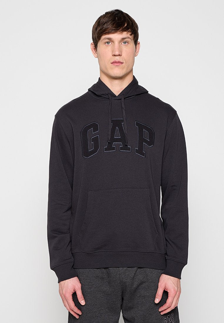 Mann trägt schwarzen GAP-Hoodie mit Fronttasche und Kordelzügen, steht vor hellem einfarbigem Hintergrund.