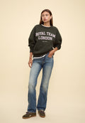 Junge Frau trägt einen dunkelgrünen Sweatshirt mit der Aufschrift "Royal Team London", blaue Jeans und olivgrüne Sneakers, steht mit der Hand in der Tasche vor einem beigen Hintergrund.