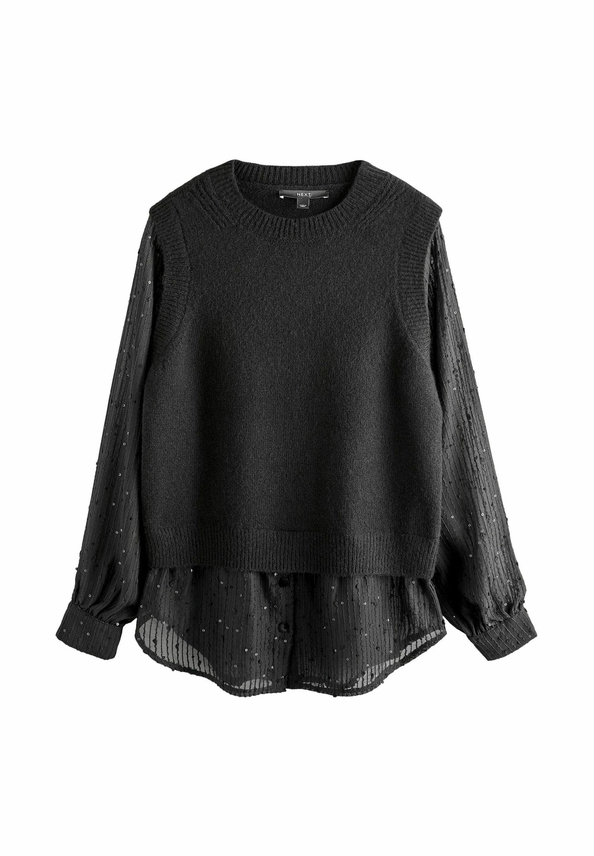 Schwarzer ärmelloser Strickpullover, der über einer transparenten schwarzen Bluse mit gepunkteter Textur getragen wird. Die Bluse hat lange, geknöpfte Ärmel.