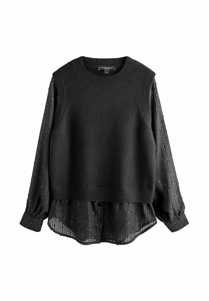 Schwarzer ärmelloser Strickpullover, der über einer transparenten schwarzen Bluse mit gepunkteter Textur getragen wird. Die Bluse hat lange, geknöpfte Ärmel.