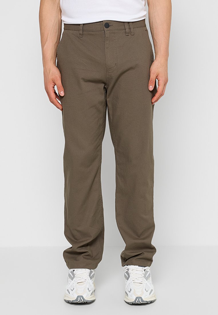 Braune Baumwollhose mit geradem Schnitt, ausgestattet mit einem Knopf- und Reißverschluss, Seitentaschen und einer strukturierten Oberfläche. Getragen mit weißen Sneakers.